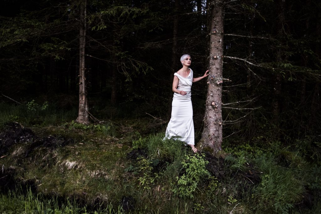 Julia Sophie Wagner in einem weißen Kleid barfuß in einem dunklen Wald stehend, mit suchendem Blick
